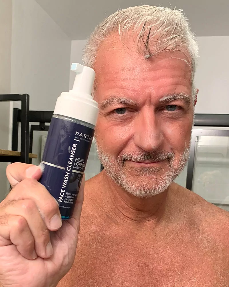 A man with gray hair holds a bottle of Particle Face Wash Cleanser.