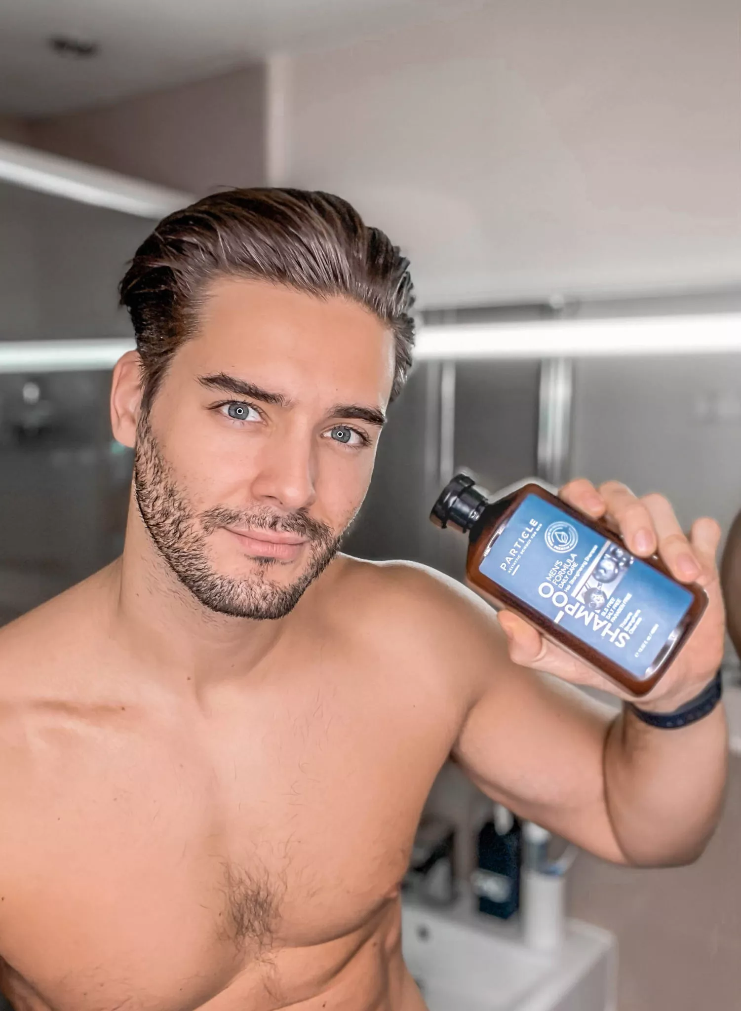 Man holding Particle Daily Care shampoo bottle, shirtless in a modern bathroom.