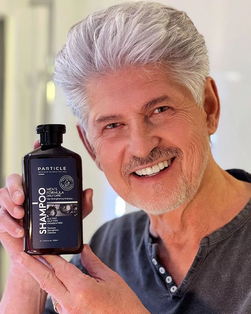 A man with gray hair and a beard smiles, holding a bottle of Particle Men's Formula Daily Care Hair Strengthening Shampoo.