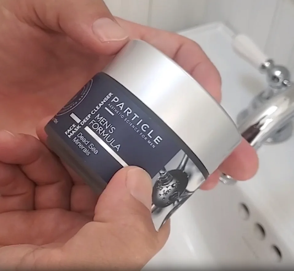 Hands holding a Particle Men's Formula face cream jar with silver lid in a bathroom